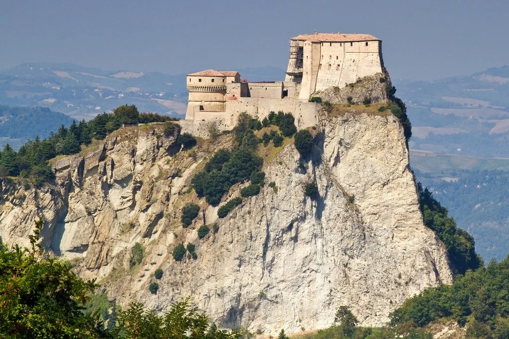 A medieval castle perched on a steep rocky cliff, overlooking a valley with rolling hills and greenery.