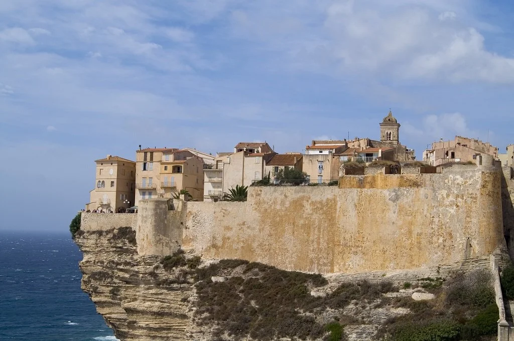 A coastal cliff town with historic buildings and a church tower overlooking the sea under a blue sky with clouds.