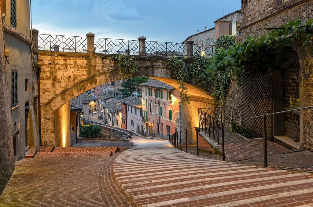 City street descending downhill with old brick buildings, a stone arch bridge, and cobblestone stairs.