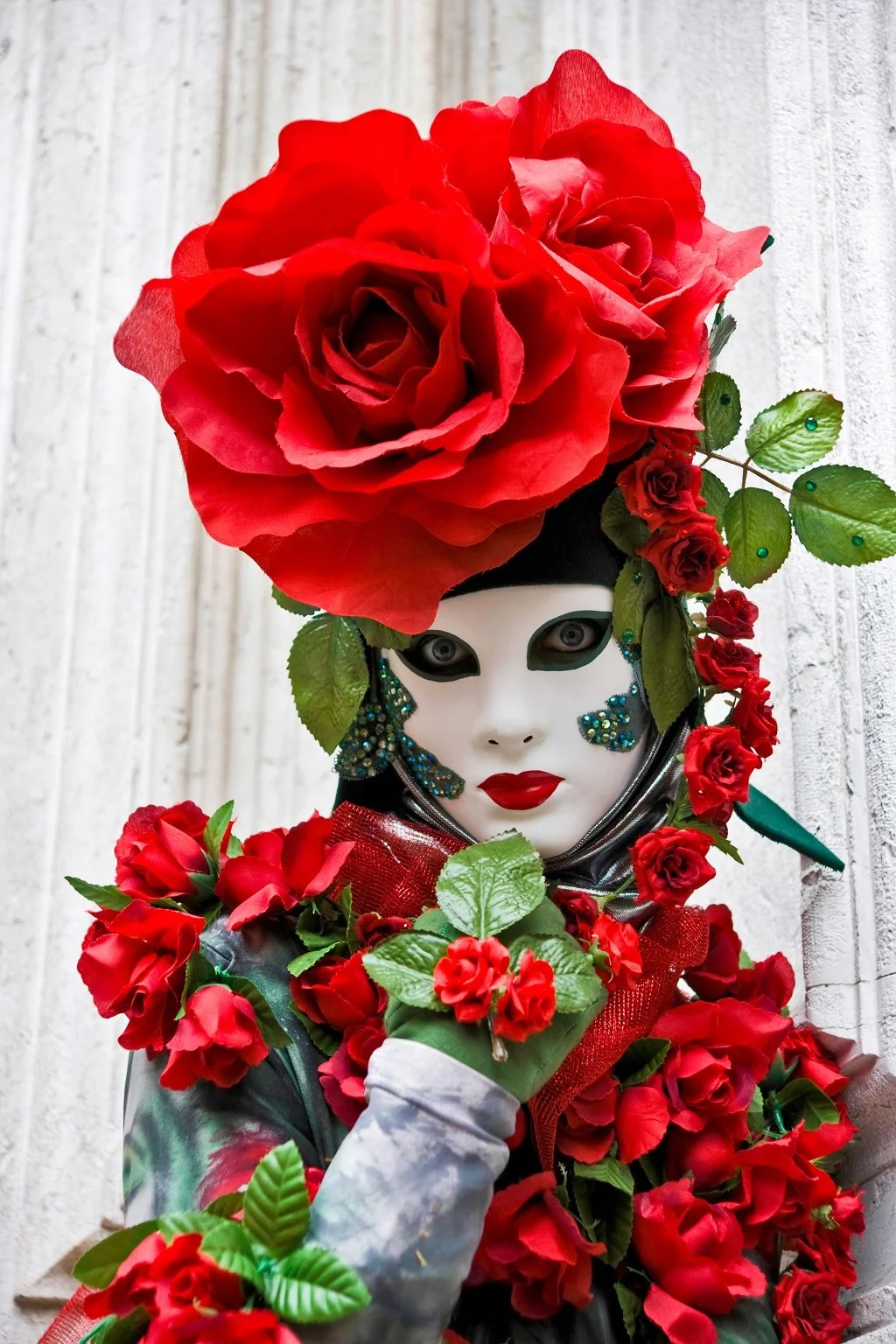 A person wearing a white mask with dark eye holes, red lipstick, and blue glitter details, surrounded by numerous red roses and green leaves, with a large rose arrangement on their head.
