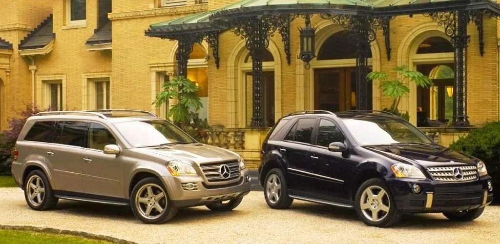 Two Mercedes-Benz SUVs parked in front of a large, elegant yellow brick house with ornate black metal awnings and green leafy trees.