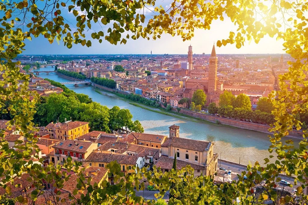 Aerial view of a city with a river running through it, surrounded by greenery and historic buildings, taken during sunset with sunlight shining from the right.