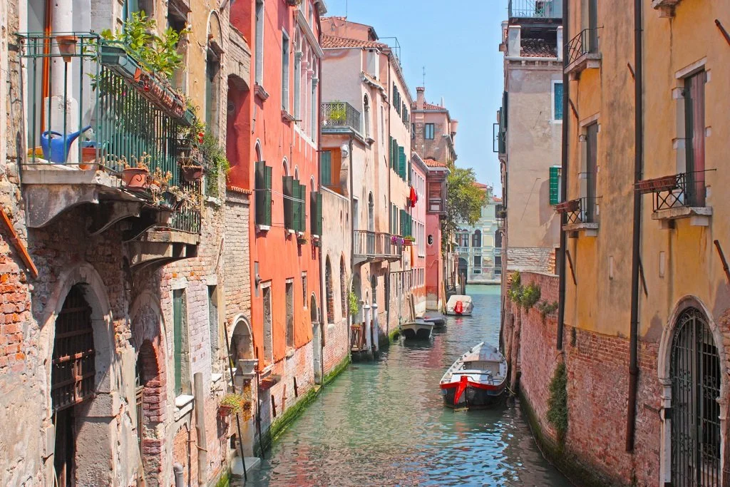 A narrow canal in Venice, Italy, with colorful old buildings on each side and boats docked along the water.
