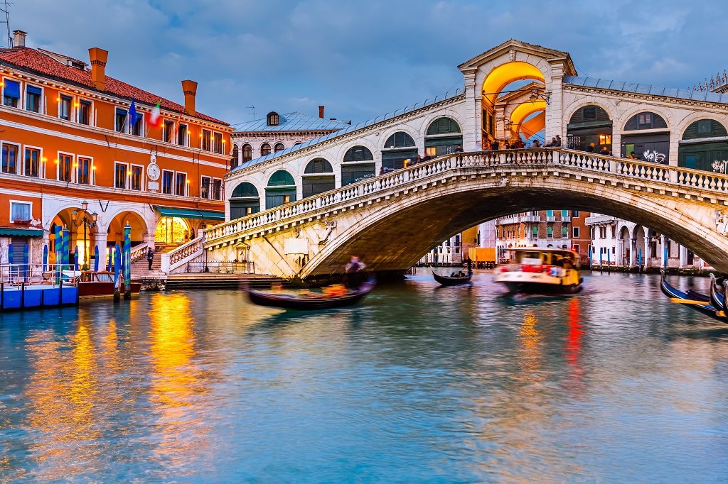 Venetian canal with boats, historic buildings, and the Rialto Bridge during sunset in Venice, Italy.