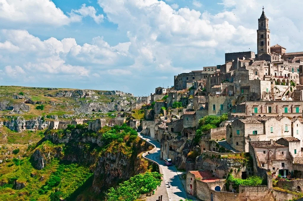Scenic view of a historic hillside town with tightly packed stone buildings, winding narrow road, and a tall church tower under a partly cloudy sky.