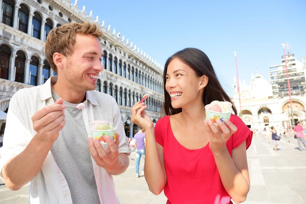 A young man and woman sharing ice cream cones and smiling at each other in a city square, with historic buildings and other people in the background.