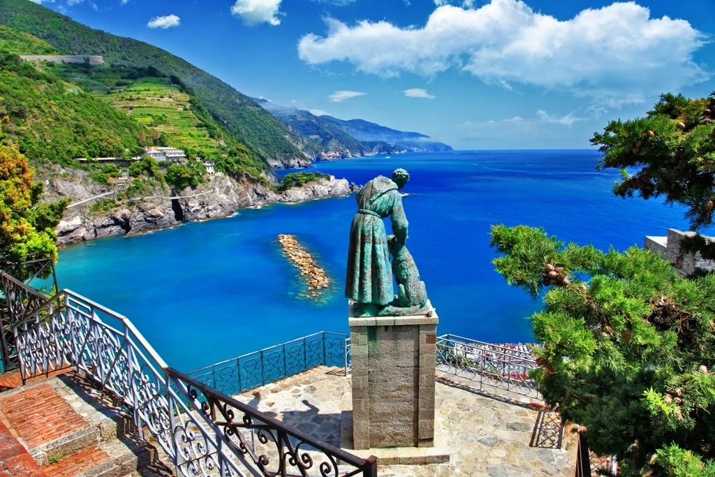 Statue overlooking a coastal scene with green hills, blue water, and a partly cloudy sky