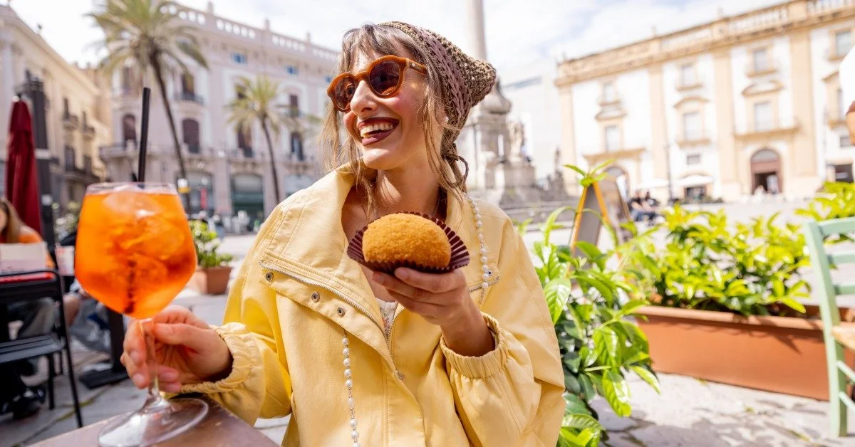 A happy woman seated outdoors in a sunny piazza at an Italian café holds a Sicilian arancino and orange spritz.