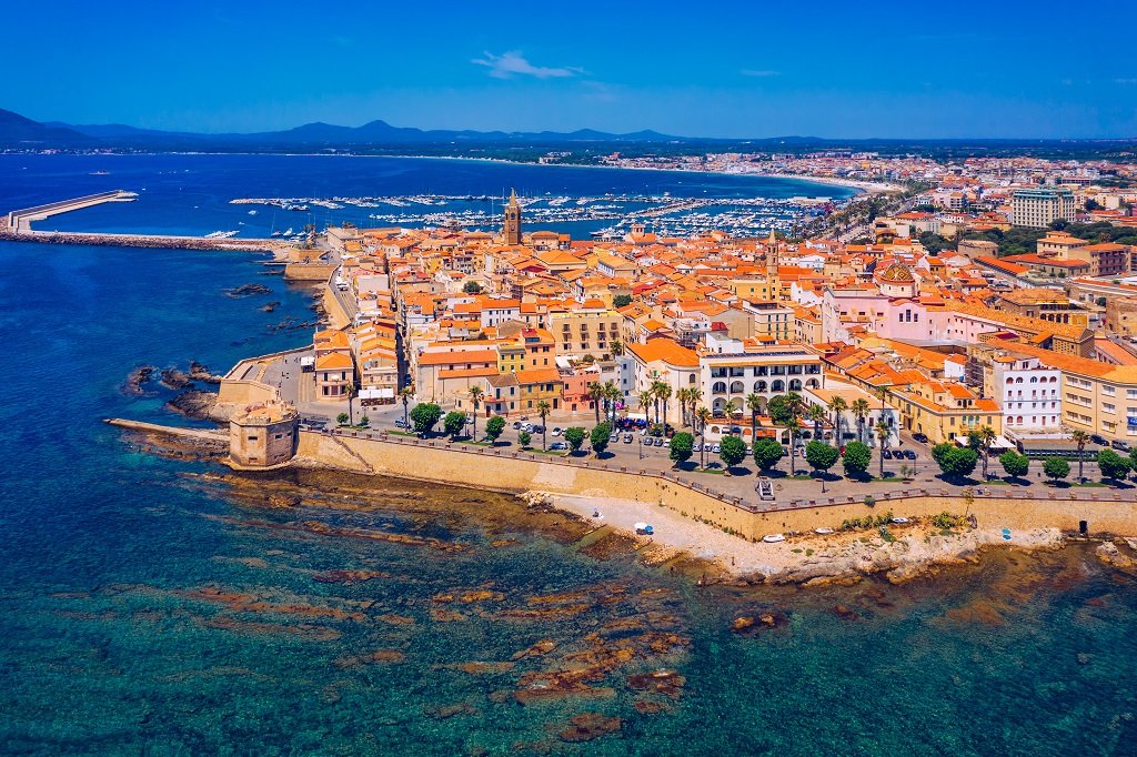 Aerial view of a coastal city with a historic harbor, orange-tiled roofs, and a marina with boats, set against a backdrop of mountains and blue sky.