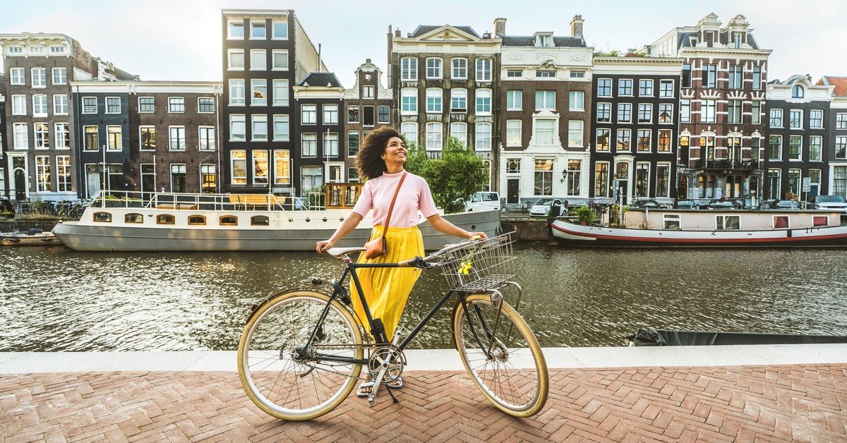 A smiling woman stands with a bicycle beside a canal, with boats and narrow row houses lining the water.