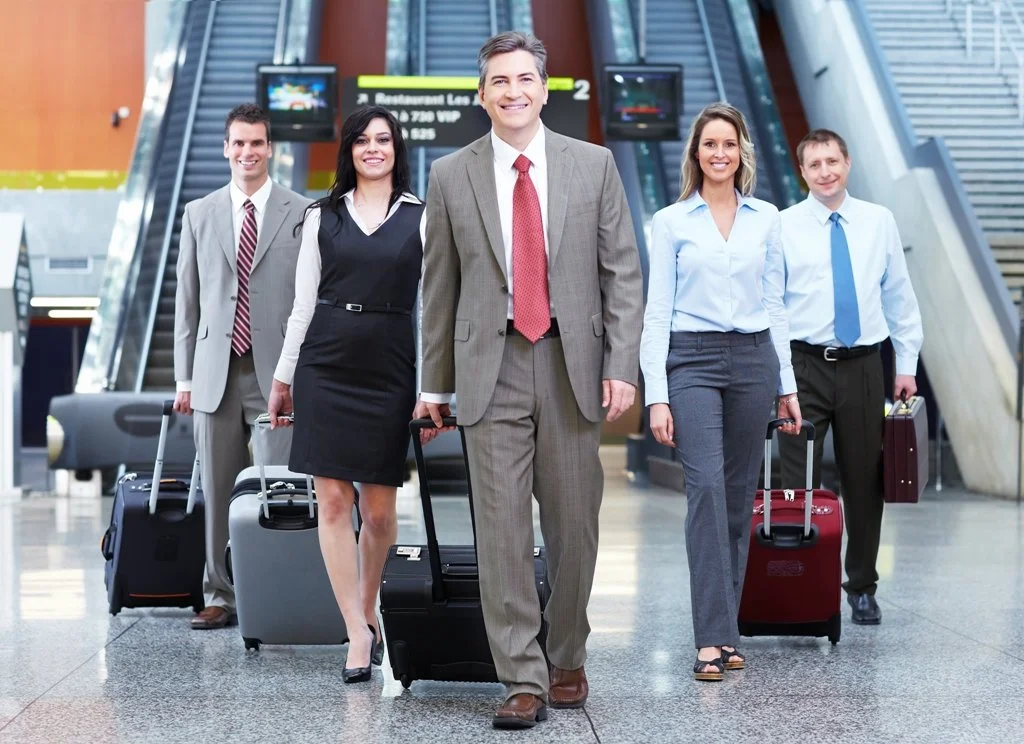 Group of five professionally dressed travelers with luggage walking through an airport.