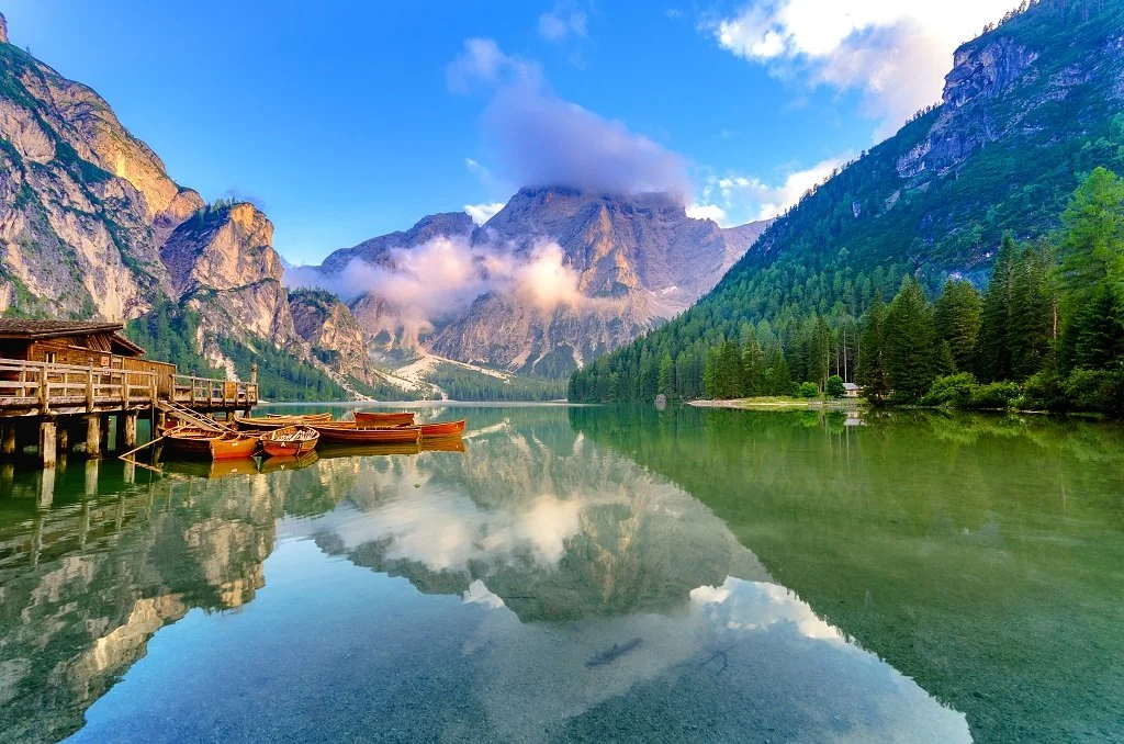 Scenic view of a mountain lake with calm water reflecting the surrounding mountains, trees, and sky, with a small wooden dock and boats on the left side.