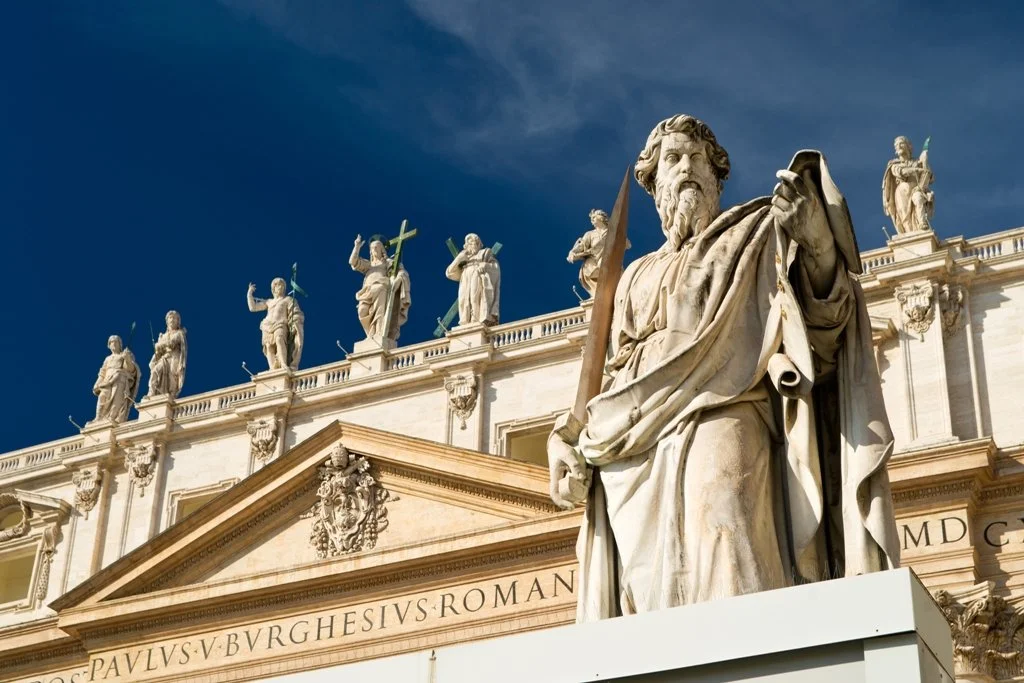 Close-up of a marble statue of a bearded man holding a sword, in front of a historical building with statues on the roof and Latin inscription.