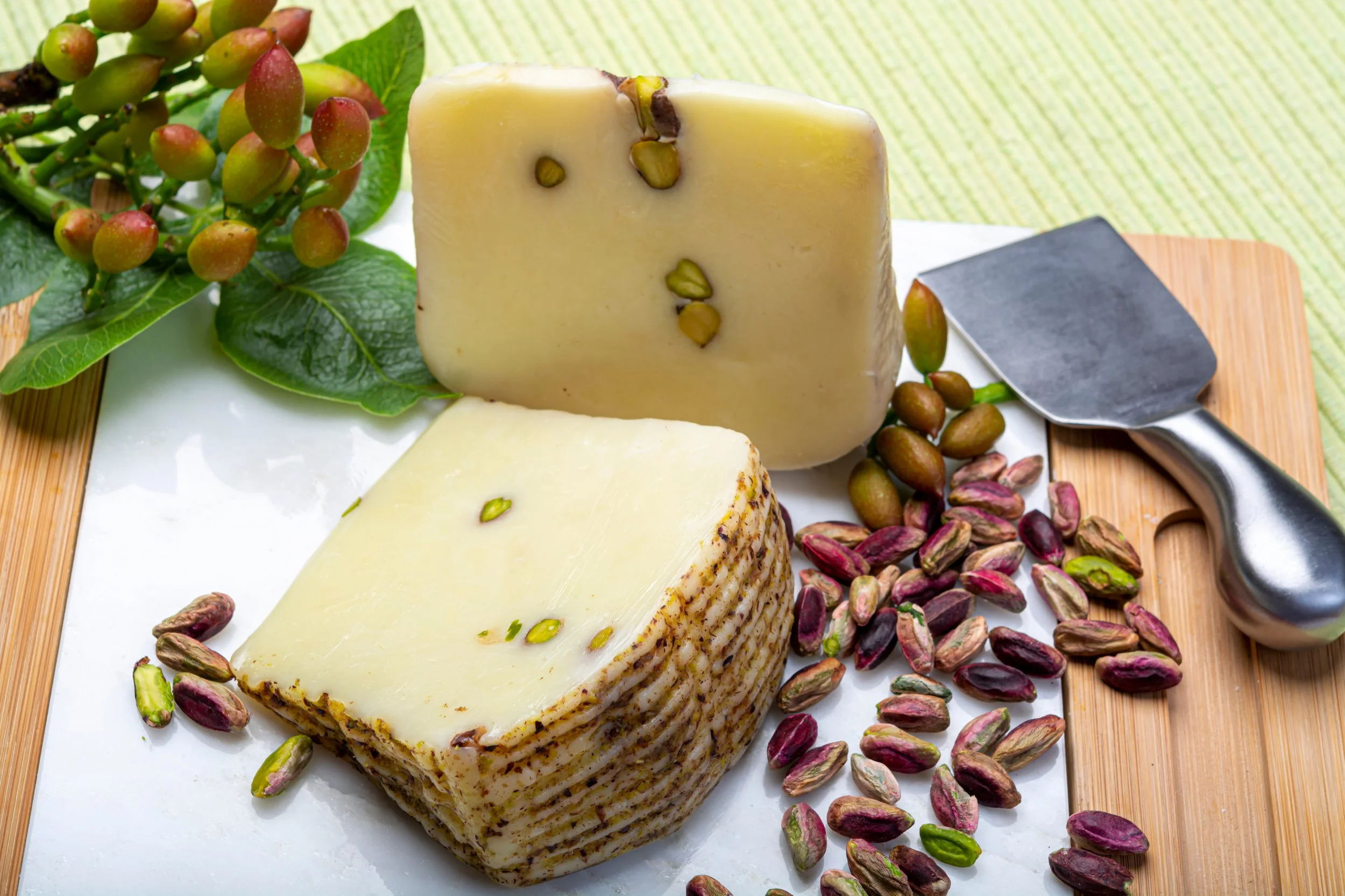 Two blocks of cheese, one with pistachio nuts, placed on a white cutting board with pistachio shells scattered around, next to a bunch of pistachio nuts and green leaves.