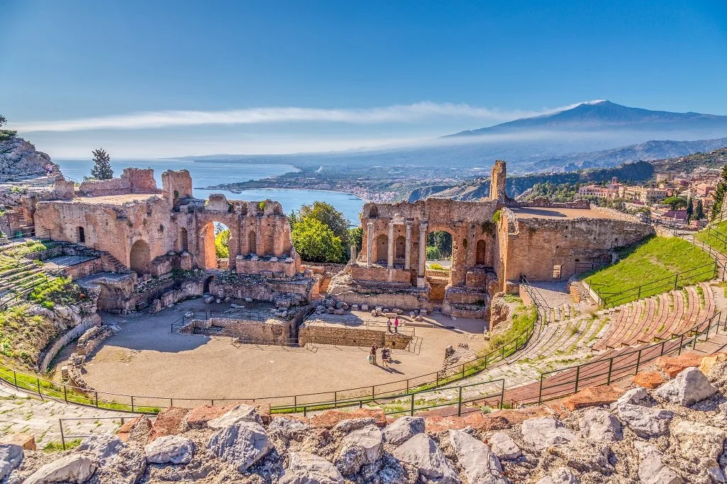 Ancient ruins of a Roman theater on a hillside with a view of a lake, mountains, and a distant volcano in the background.