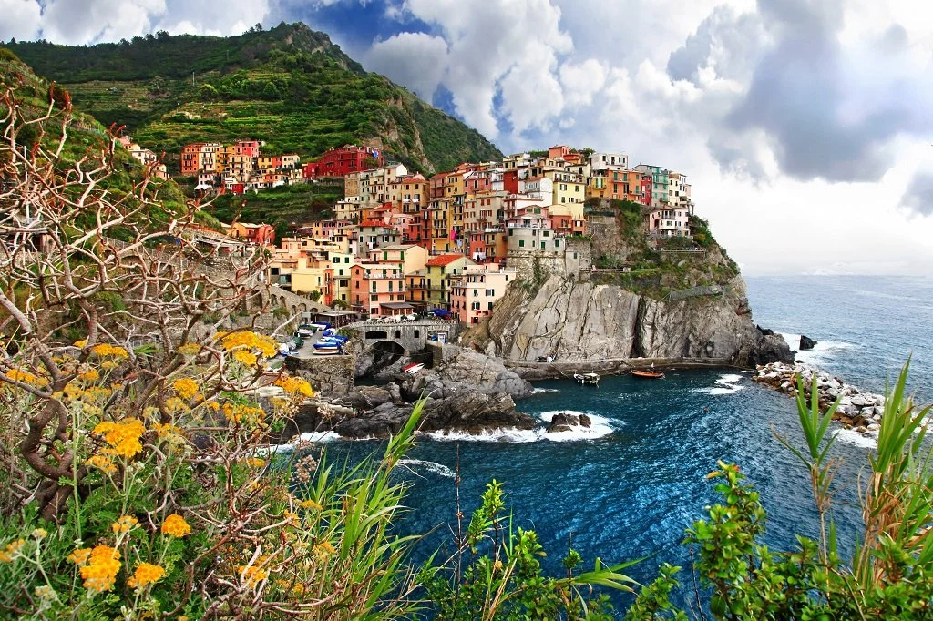 Colorful houses climbing a steep hillside above a rocky coastal cove with boats and blue water, surrounded by lush green vegetation and cloudy sky.