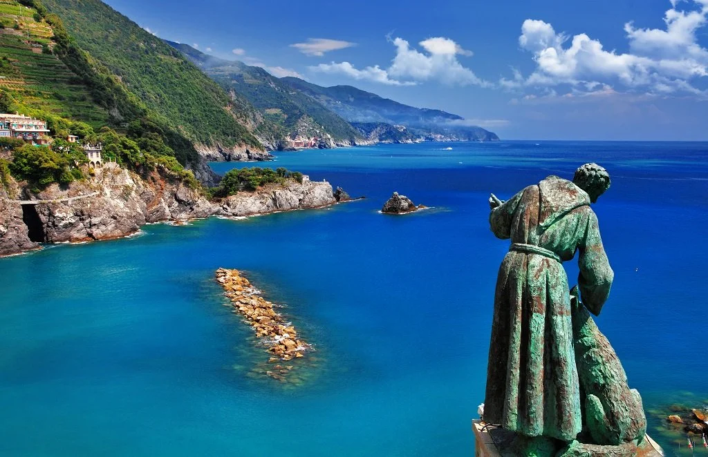 Bronze statue of a person looking over the coastline of a blue sea with green hills and a partly cloudy sky