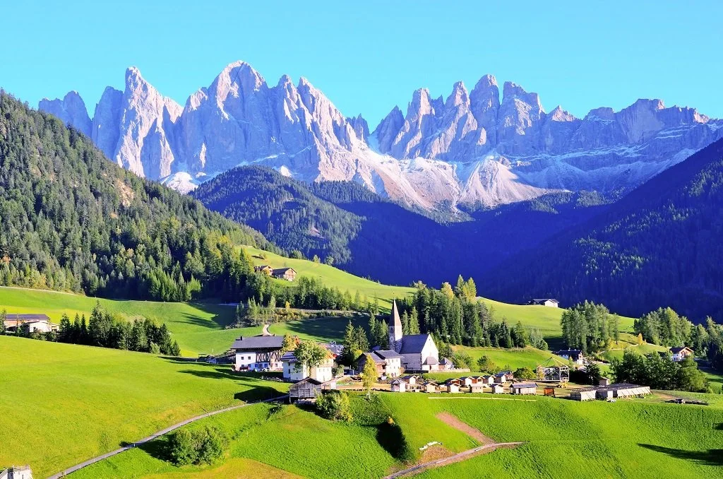Scenic view of a mountain landscape with rugged peaks, lush green hills, and a small village with a church in the foreground.