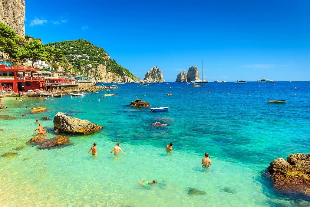 People swimming and relaxing in a clear blue ocean near a rocky shoreline with colorful buildings, green hillside, and several sailboats and rocks in the distance.