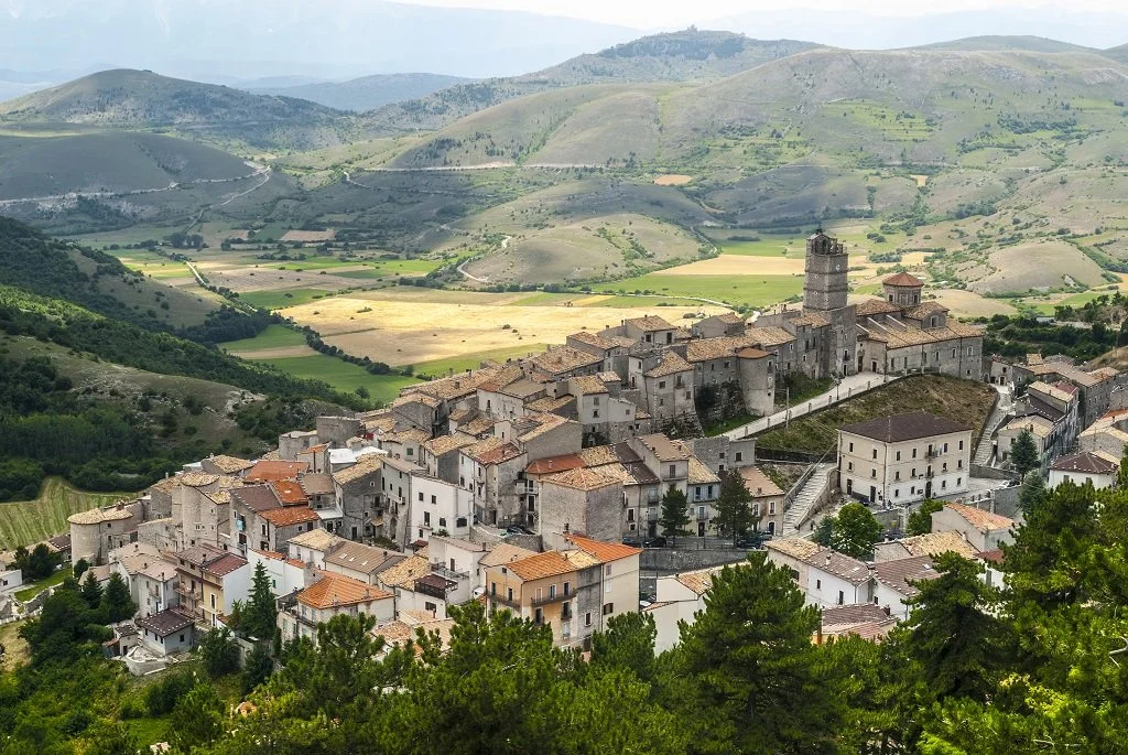 A historic Italian hilltop town with stone buildings and a prominent tower, surrounded by lush green hills and fields under a partly cloudy sky.