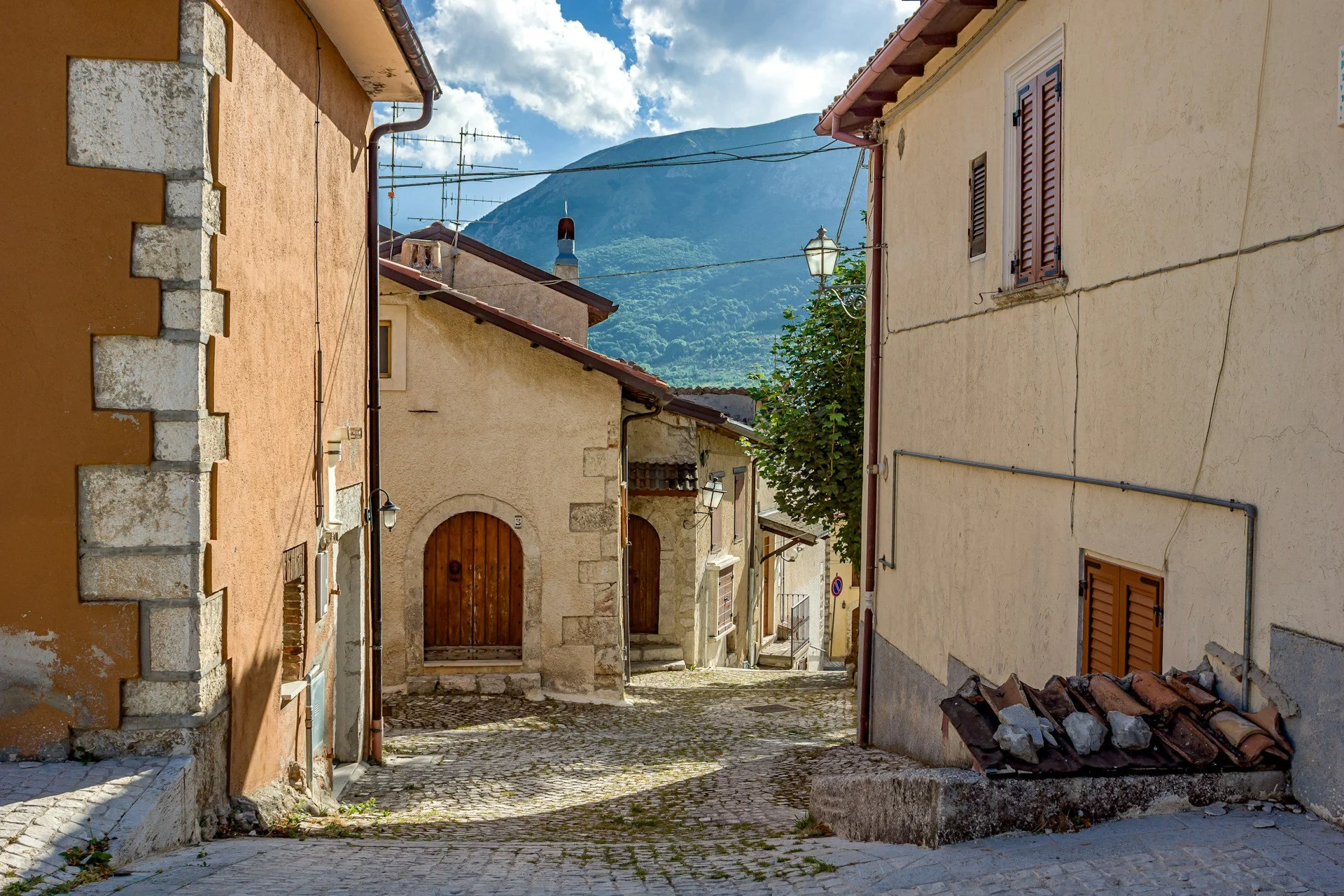 A narrow, sloping cobblestone street framed by historic stone and stucco buildings in the medieval mountain village of Calascio, located in the Province of L'Aquila within the Abruzzo region of Italy.
