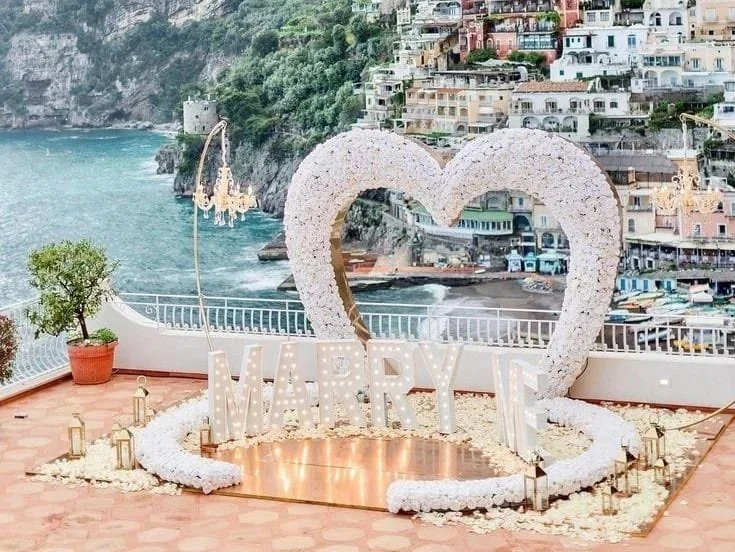 Decorative wedding setup with a large heart-shaped frame covered in white flowers, illuminated letters spelling 'MARRY ME,' candles, and flower petals on the ground, overlooking the sea with a cliffside town in the background.