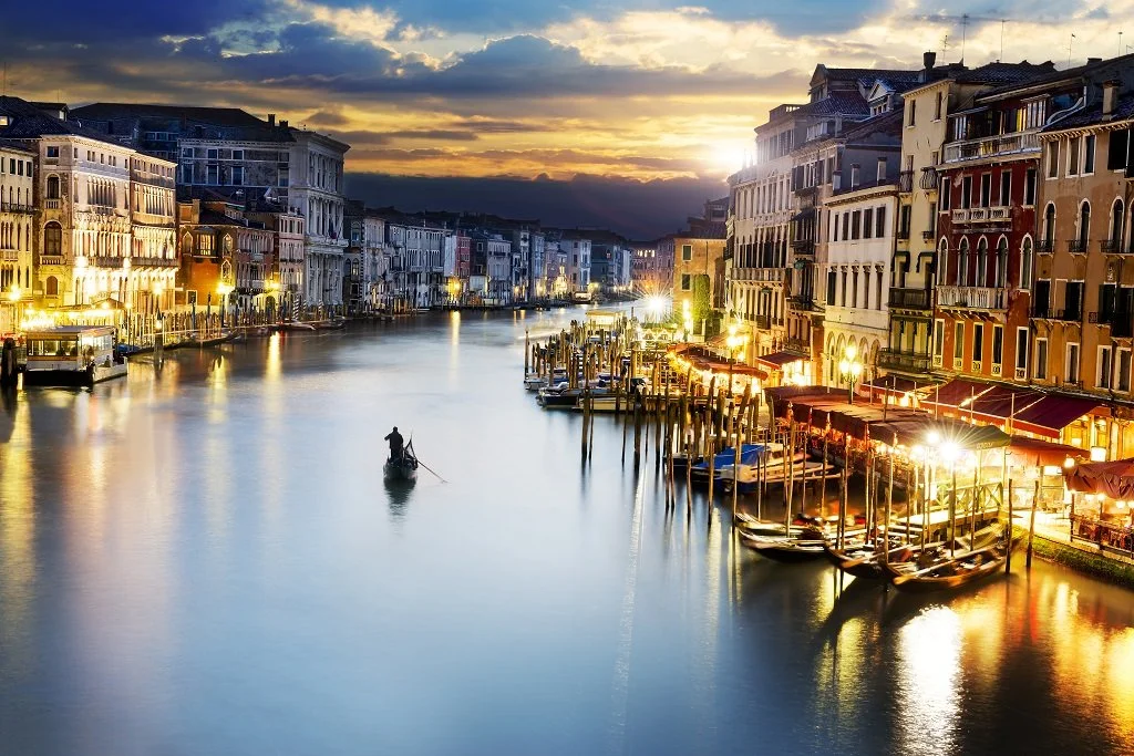 Venetian canal at sunset with boats docked along the sides and a gondolier in a gondola on the water.