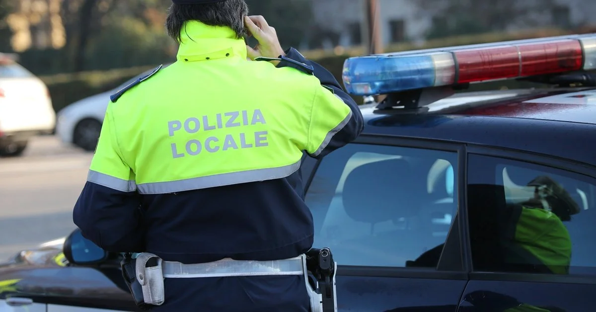 An Italian Polizia Locale stands facing their vehicle on the phone. They wear a hi-vis yellow safety coat and a hat.
