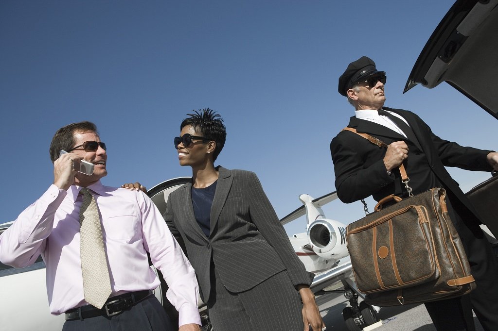 Three businesspeople smiling and talking outside of a private jet, with one man using a cell phone, an airplane in the background, and a clear blue sky.
