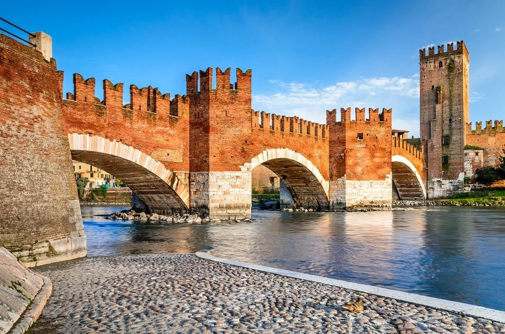 Medieval red brick castle with crenelated walls and towers over a river, with a cobblestone path in the foreground and a partly cloudy sky.