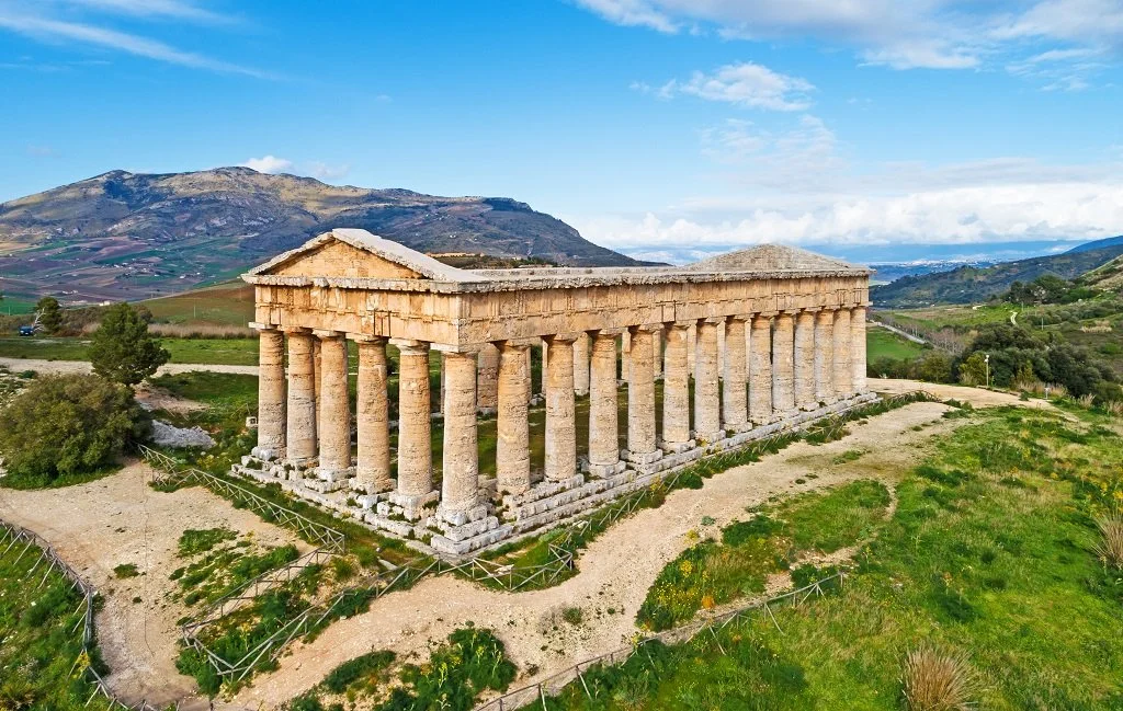 Ancient Greek temple with tall columns in a lush landscape, mountains in the background, under a partly cloudy sky.