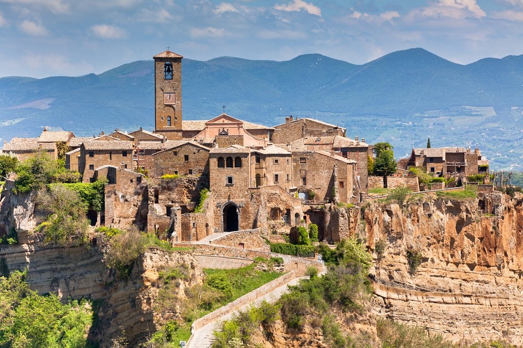 A historic village perched on a cliff with stone buildings and a church tower, with mountains in the background.