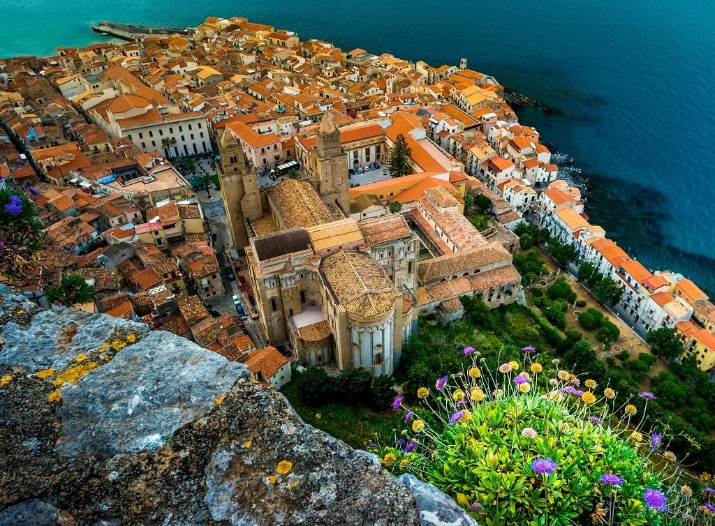 Aerial view of a coastal town with orange-tiled roofs, historic church, and surrounding greenery, overlooking the ocean.