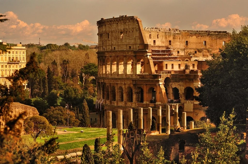 The Colosseum, an ancient Roman amphitheater, during sunset in Rome, Italy, surrounded by trees and city buildings.