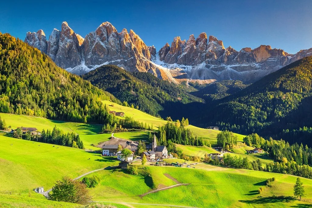 Scenic view of green rolling hills, scattered houses, and a small church in a valley with a backdrop of tall, snow-capped mountains under a clear blue sky.