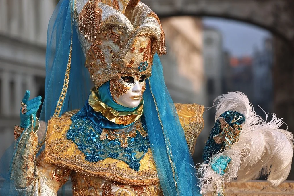 A person dressed in a Venetian-style carnival costume with gold, blue, and white ornate fabric, a gold and silver mask, and a tall, elaborate headdress. They hold a feathered fan.