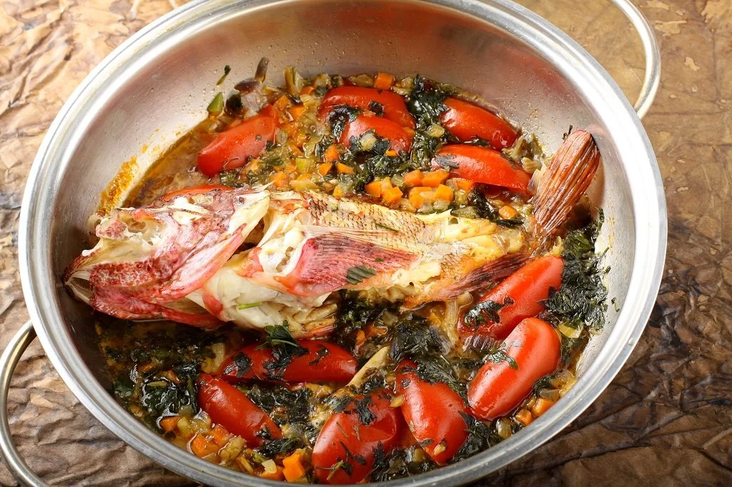 A pot containing a cooked fish with vegetables including chopped tomatoes, carrots, and leafy greens.