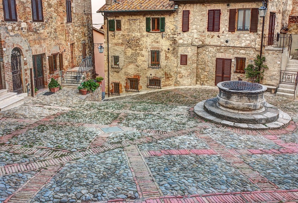 Old European cobblestone square with a stone well at the center surrounded by historic buildings with wooden shutters and potted plants.