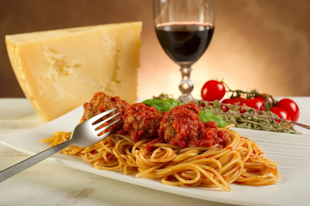 Spaghetti with meatballs, basil, and tomato sauce on a white plate, with cheese, cherry tomatoes, and a glass of red wine in the background.