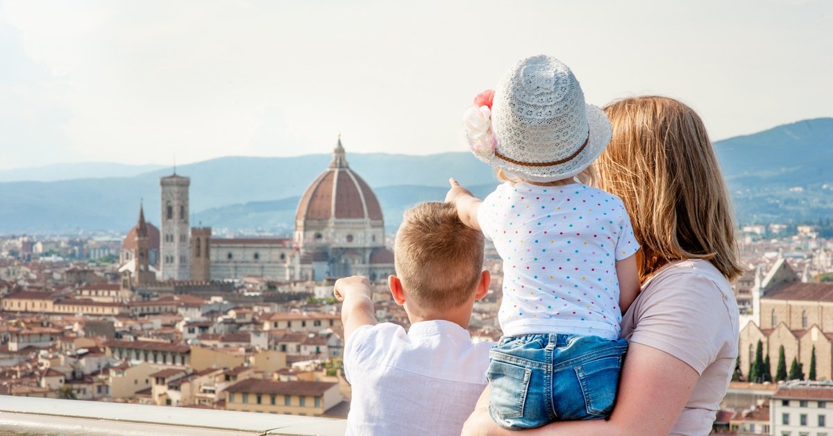 A mother holds a toddler, and a small child stands next to them. They're on a balcony pointing to the city below.