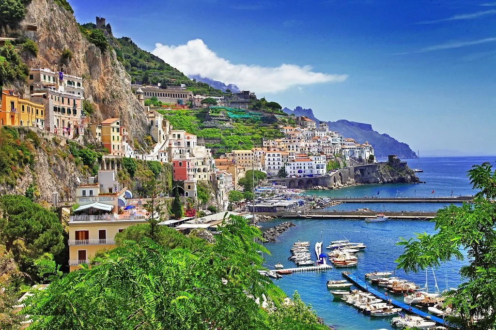 Colorful hillside buildings overlooking a harbor with boats and yachts in a coastal town, with lush greenery in the foreground and a blue sky.