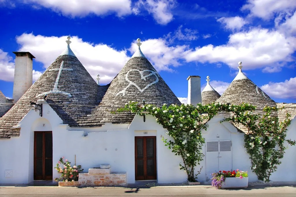 White house with conical roofs, graffiti of a heart with a heartbeat line and a peace sign, among green vines and colorful flowers, against a bright blue sky with scattered clouds.