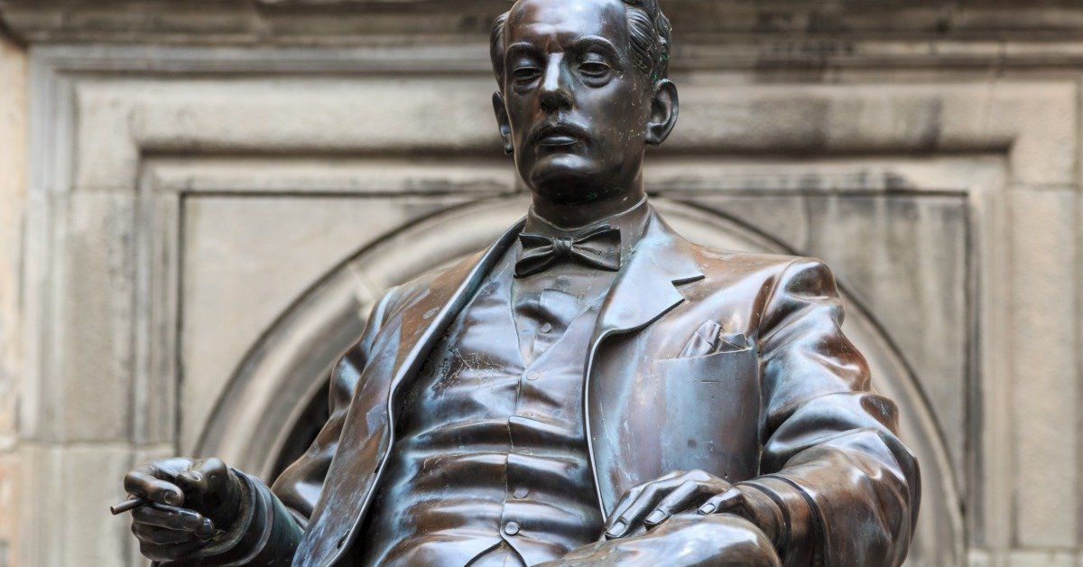 A bronze statue of composer Giacomo Puccini standing with a coat and hat in a public square with buildings behind.