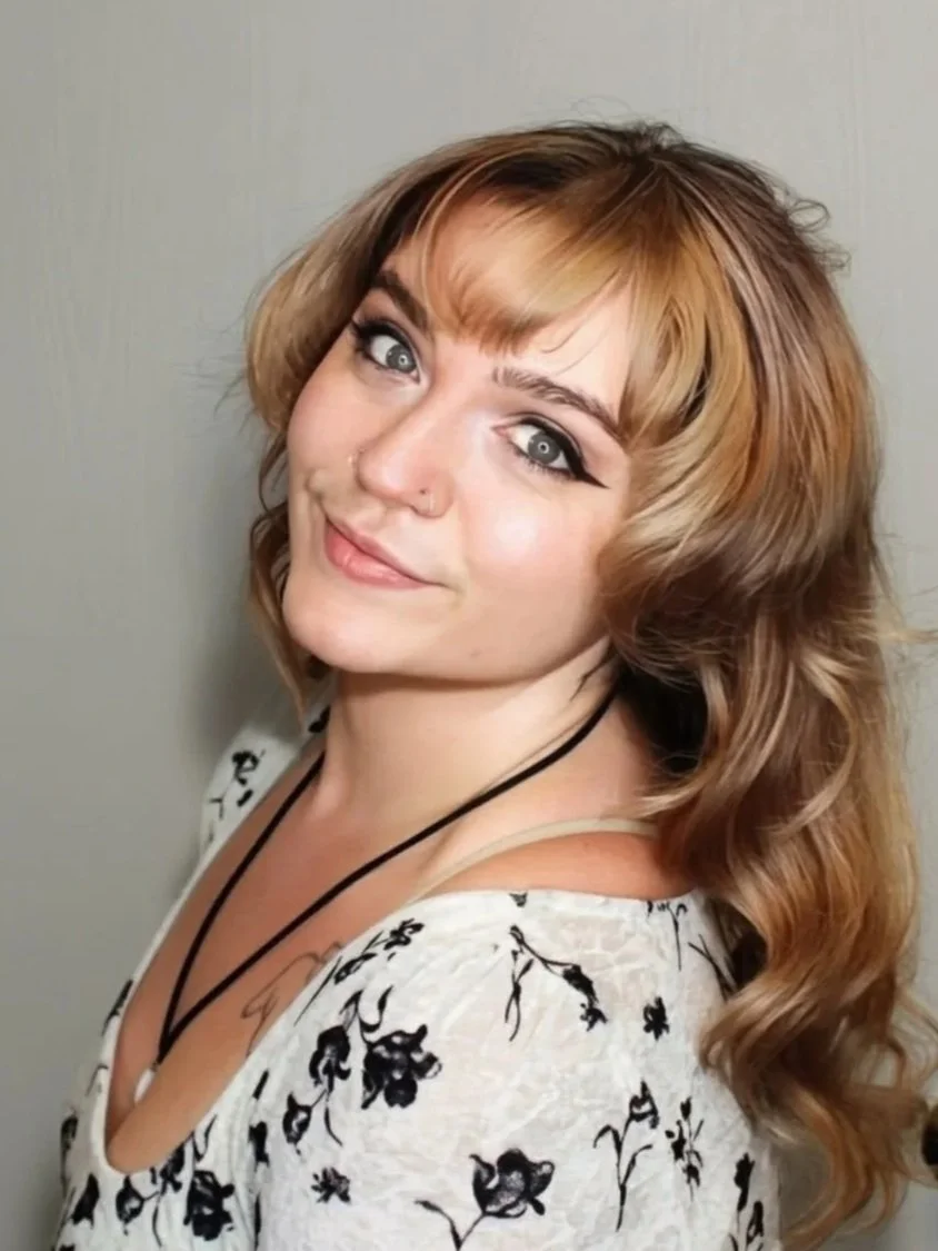 Close-up of a woman with long, wavy blonde hair, blue eyes, and a nose piercing, smiling softly against a dark gray background.
