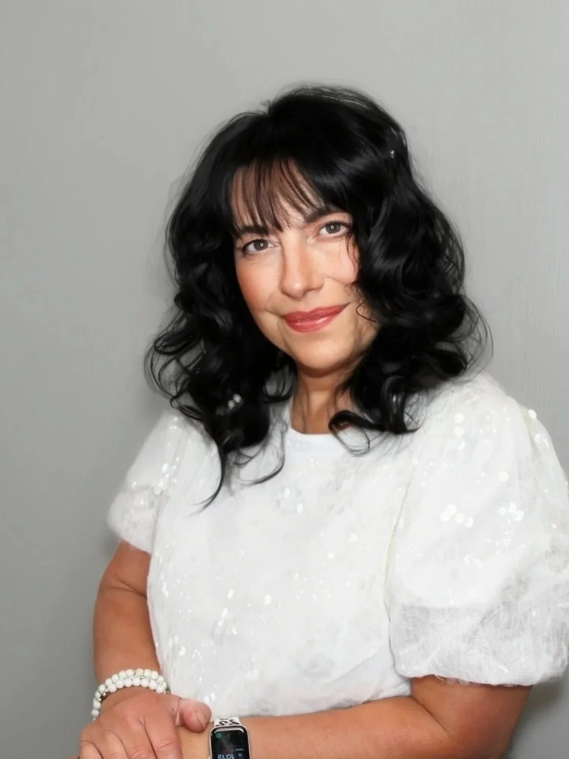 A woman with black hair, wearing a pink top, smiling with her hand touching her face, against a gray background.