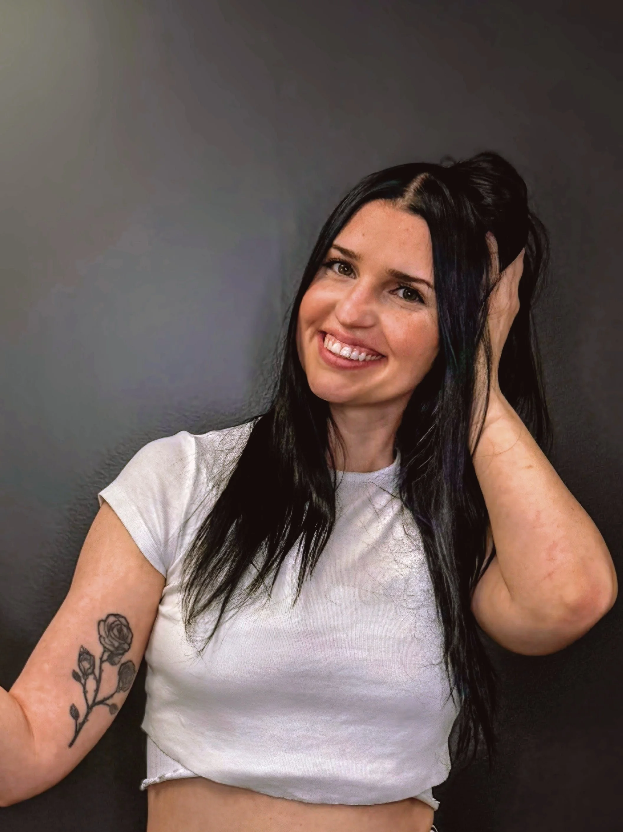 A woman with long black hair, smiling, wearing a white t-shirt with a tattoo of a flower on her left arm, posing against a dark gray background.