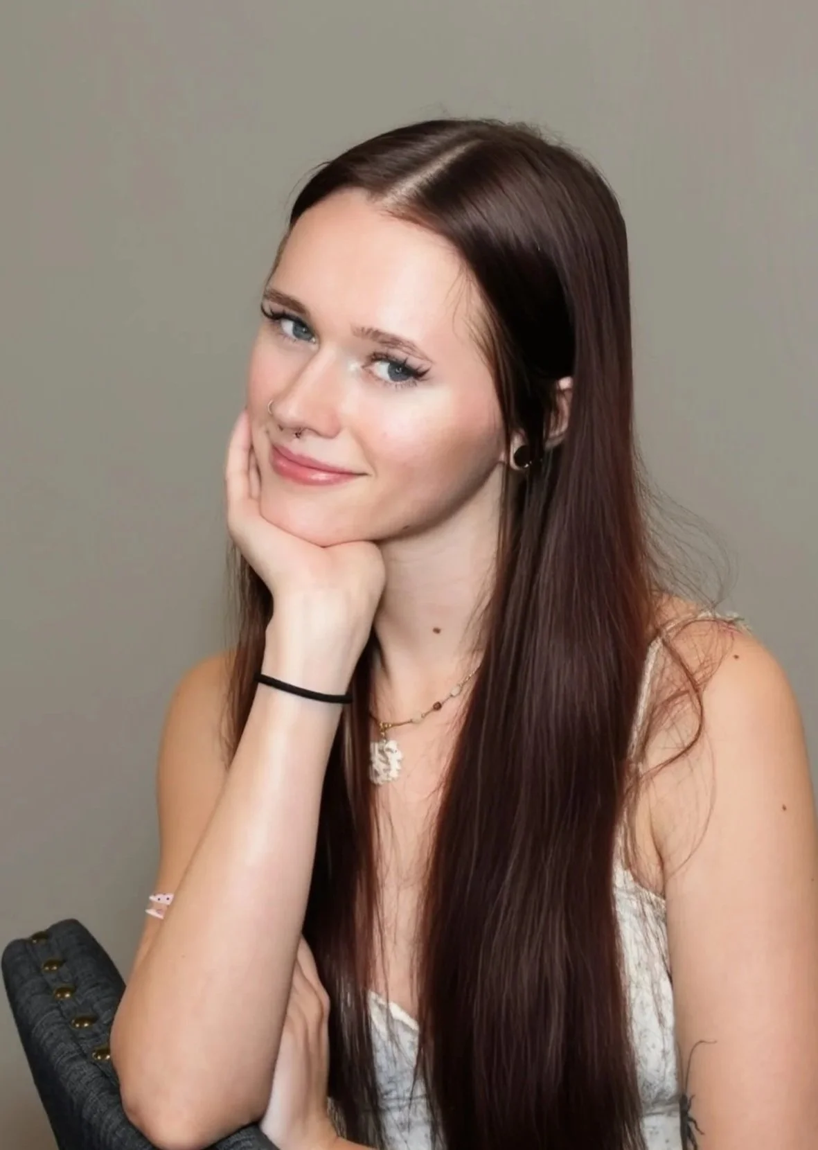 Portrait of a young woman with long, curly red hair, wearing a pink knitted sweater, black earrings, and a necklace with a pendant, smiling at the camera against a plain dark grey background.
