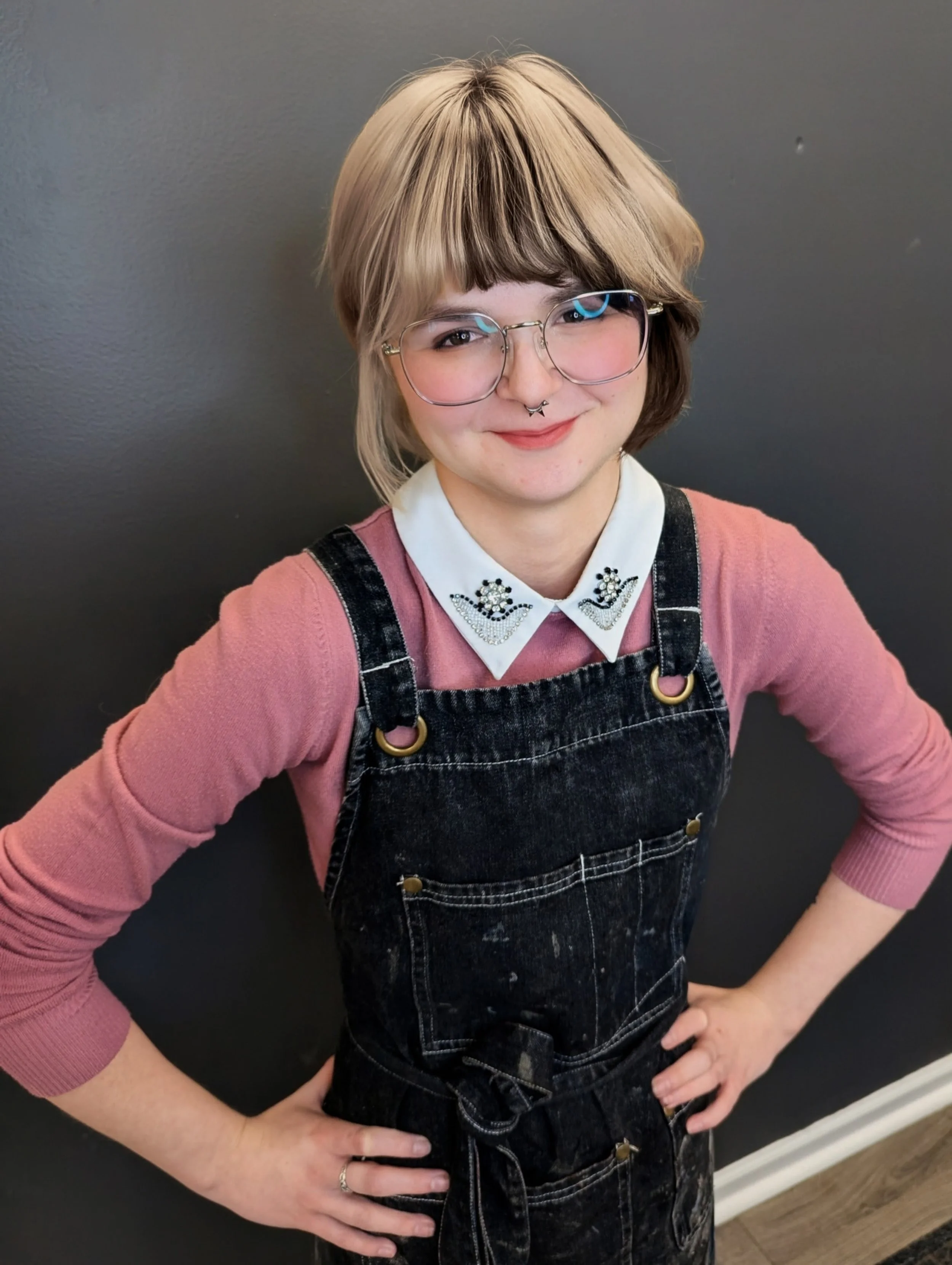 A woman with short, blonde hair wearing glasses, a white collared shirt with decorative embroidery, a pink sweater, and denim overalls, standing with her hands on her hips in front of a dark wall.