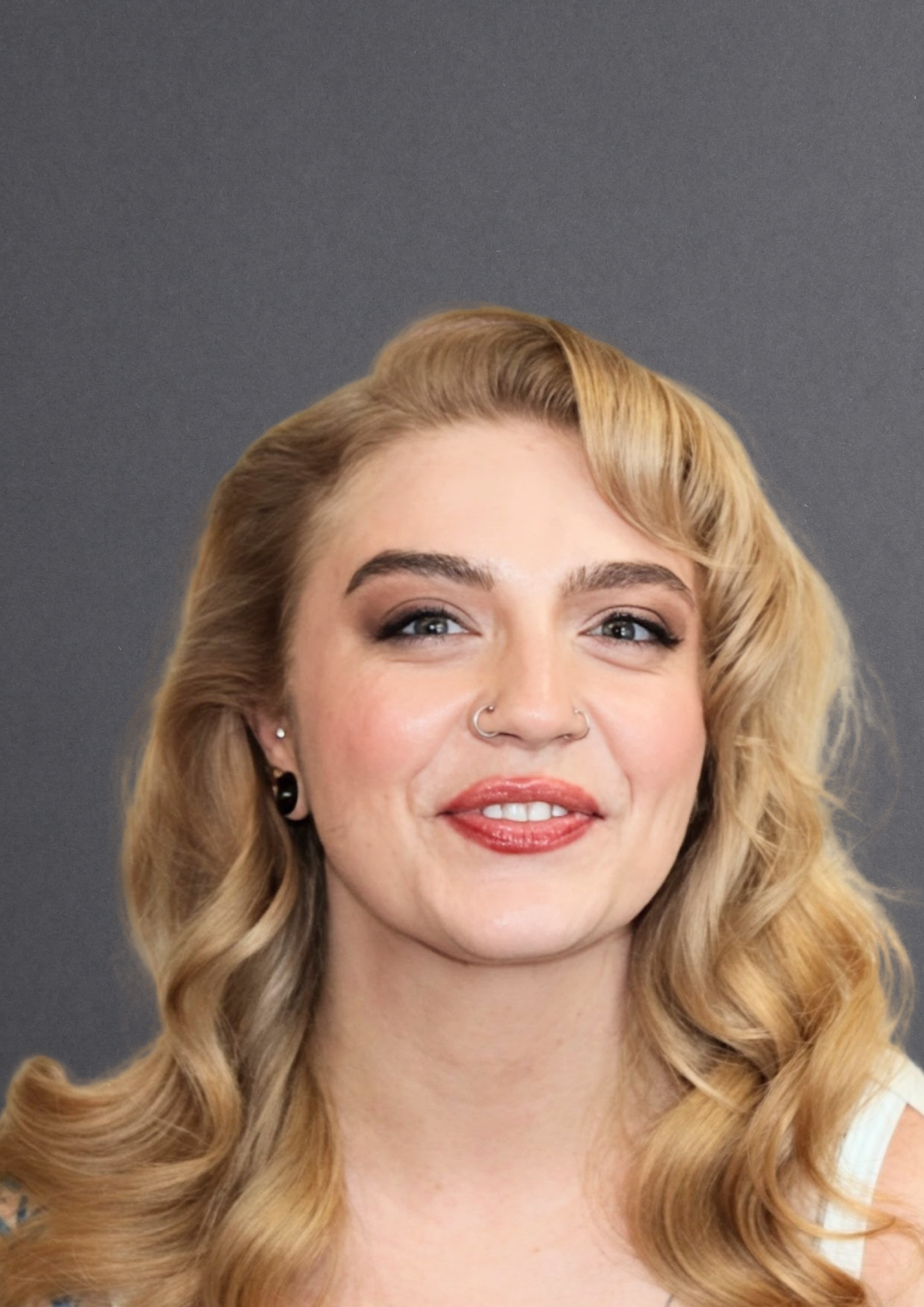 Close-up of a woman with long, wavy blonde hair, blue eyes, and a nose piercing, smiling softly against a dark gray background.