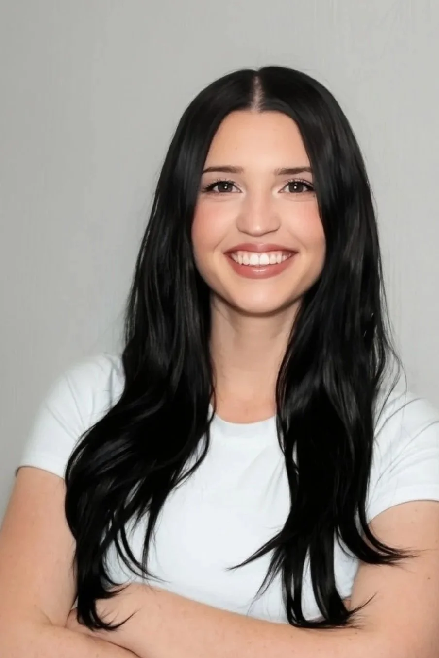 A woman with long black hair, smiling, wearing a white t-shirt with a tattoo of a flower on her left arm, posing against a dark gray background.